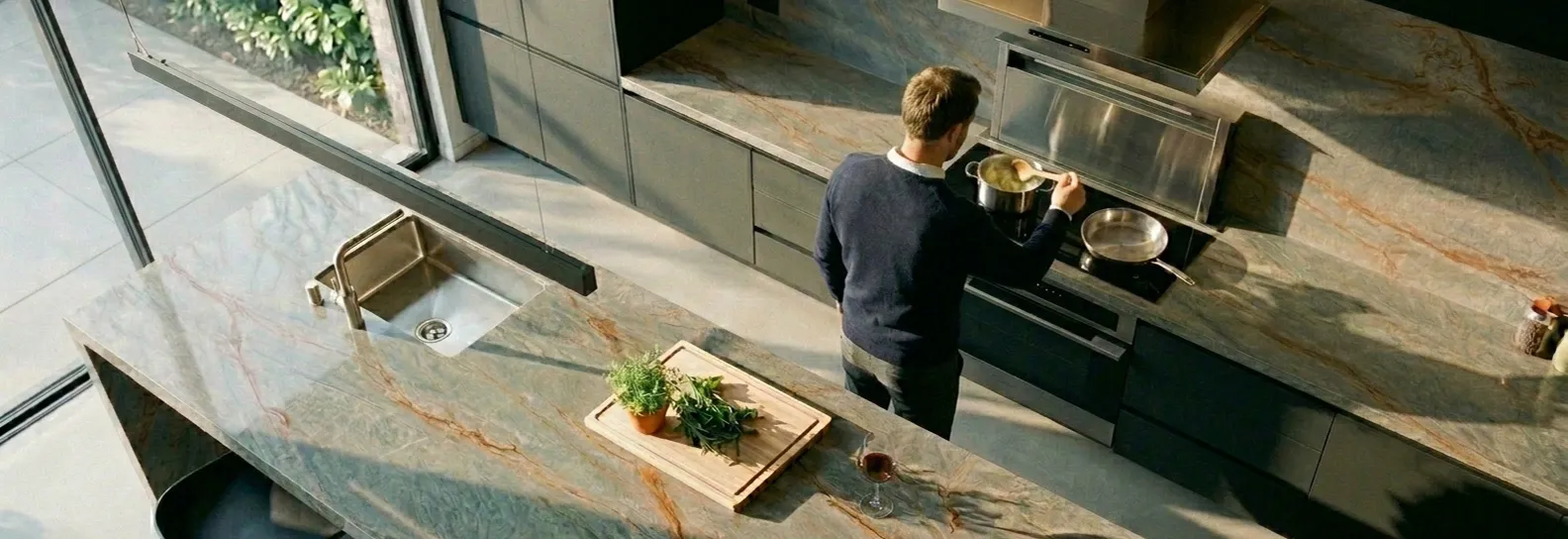 Espectacular mesón de piedra sinterizada gris con vetas doradas en una cocina de lujo iluminada por el sol, donde un hombre cocina con total confianza y libertad.